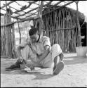 Man wearing Western clothes sitting outside the expedition hut, eating from an enamel dish