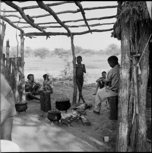 /Ti!kay sitting with other people in the expedition hut structure, with cooking pots and a jerry can on the ground near them