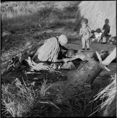 /Ti!kay lying down during a curing ceremony, with a woman aiding him, a child standing next to him