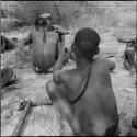 Man squatting, checking his arrows for balance, view from behind, with arrows and a quiver on a log near him