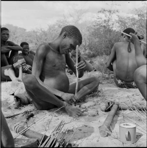 Man sitting, rolling a fire stick, with a quiver and an enamel cup on the ground next to him, arrows drying on logs in front of him