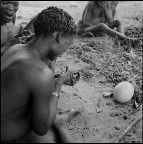 Man filing an arrow shaft, sitting with a group of men, an ostrich eggshell on the ground in front of him