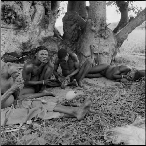 Group of men at the base of a baobab, some working on arrows, some sleeping