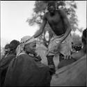 /Gunda dancing at a daytime dance, next to a woman wearing a headdress, sitting with a group of women