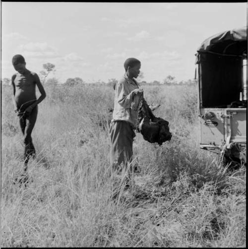 Man carrying a large piece of meat to the expedition Jeep, with another man walking near him