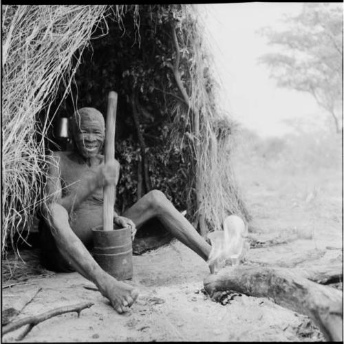 Man sitting next to a fire in front of his skerm, pounding with a mortar and pestle