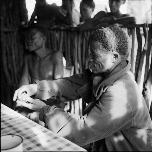 ≠Toma wearing a coat, holding a beetle in his hand, sitting with /Ti!kay at a table in the expedition tent, helping expedition members to identify insects, with people standing outside behind them