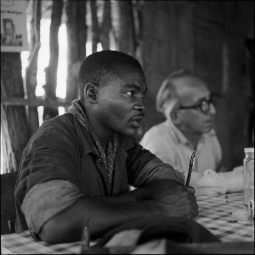 Kernel Ledimo and Charles Koch sitting at a table in the expedition tent, talking with /Ti!kay and ≠Toma who are helping expedition members to identify insects