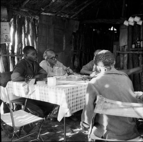 /Ti!kay and ≠Toma sitting at a table with Laurence Marshall and other expedition members, working with them to identify insects