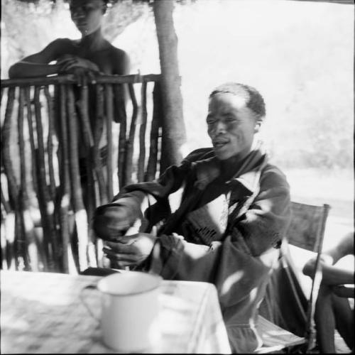 ≠Toma sitting at a table in the expedition tent, talking, helping expedition members to identify insects, with a man standing outside behind him