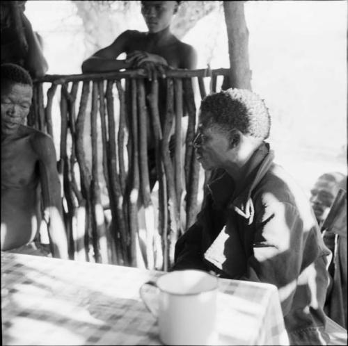 ≠Toma and /Ti!kay sitting at a table in the expedition tent, helping expedition members to identify insects, with people standing outside behind them