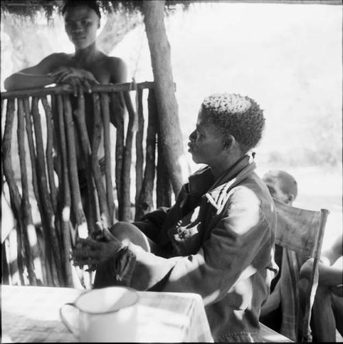 ≠Toma wearing a coat, sitting at a table in the expedition tent, helping expedition members to identify insects, with a man standing outside near him