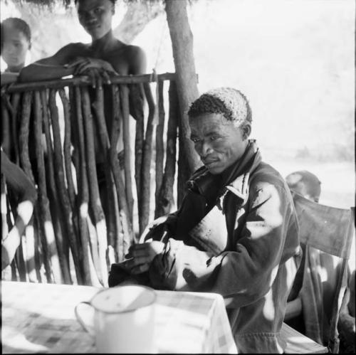 ≠Toma wearing a coat, sitting at a table in the expedition tent, helping expedition members to identify insects, with two people standing outside near him