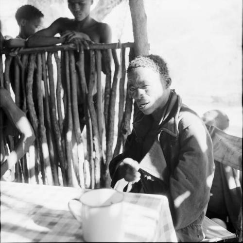 ≠Toma wearing a coat, sitting at a table in the expedition tent, helping expedition members to identify insects, with two people standing outside near him