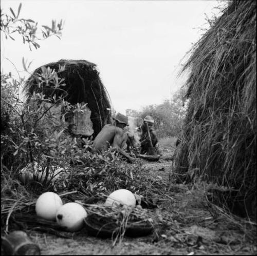 /Ti!kay sitting with a woman pounding with a mortar and pestle, with a boy sitting in a shelter skerm behind them, animal skin hanging in the opening of the skerm, ostrich eggshells and a melon on the ground near them