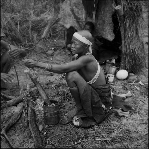 Woman using a mortar and pestle, with /Ti!kay's hands putting grass on a fire next to her, boy sitting in a shelter skerm behind her