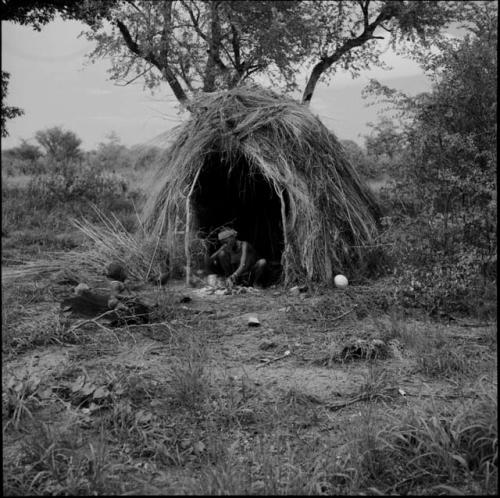 Woman squatting in the entrance of her skerm, putting sticks on the fire, with her baby sitting on a hide in front of the skerm