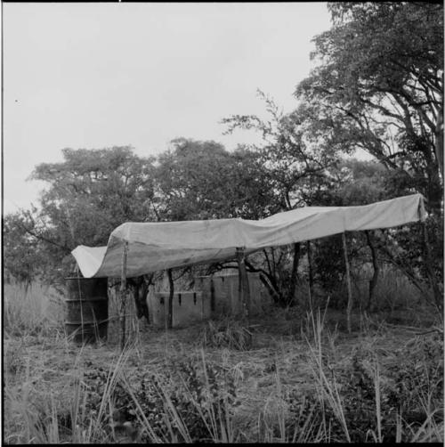 Expedition rain-saver constructed with canvas stretched across four posts, higher in the back, with a barrel at the lower end to catch the water, view from the side