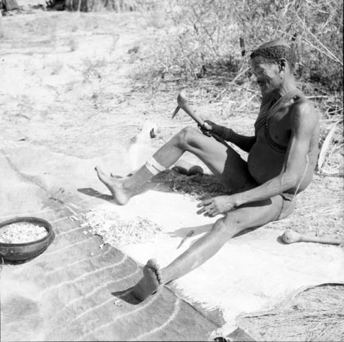 Man sitting on a hide, scraping it with an adze, with the scrapings in a bowl and on the hide in front of him