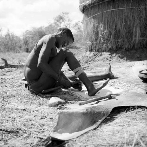 Man scraping a hide, with his adze lying next to him on the ground