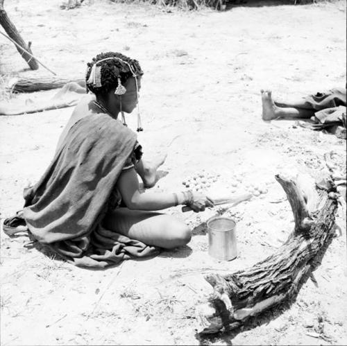 Woman cooking mangetti nuts in the ashes of a fire, pushing them with a fire paddle