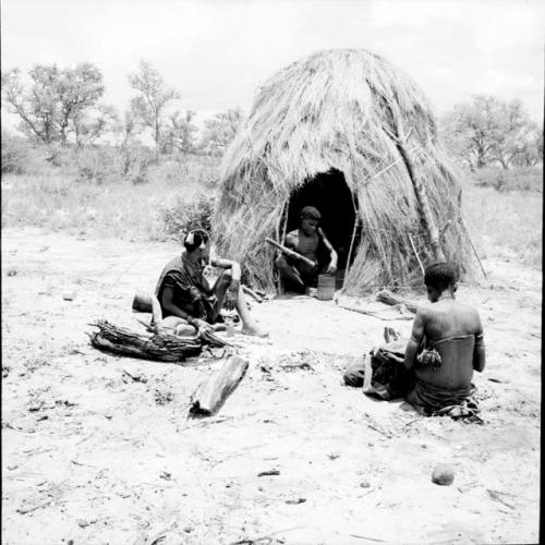 Man pounding with a mortar and pestle, sitting in the opening of his skerm, with two women preparing mangetti nuts near him