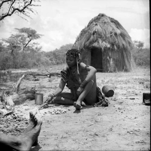 Woman cooking mangetti nuts, turning them with a fire paddle, with a skerm in the background