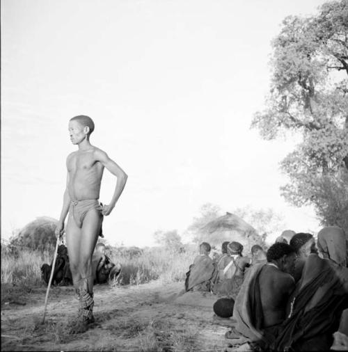 Man dancing, with two groups of people sitting near him, skerms in the background