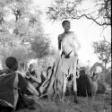 Woman wearing a kaross, holding a blanket, standing next to a group of people sitting