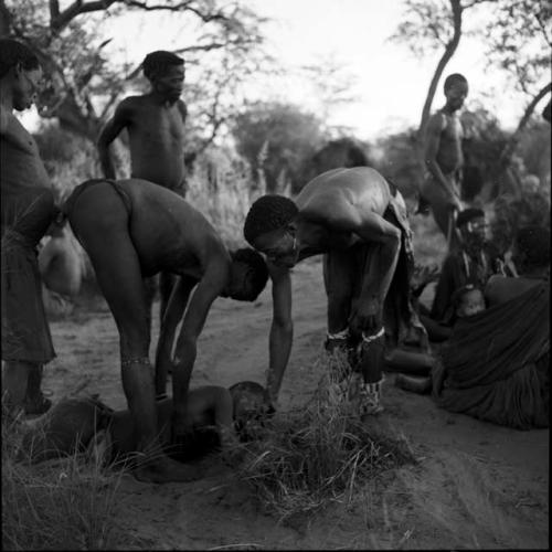 /Ti!kay and another medicine man leaning over, tending to a man lying on the ground in trance, with people sitting and standing in the background