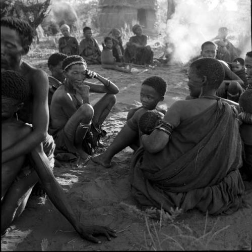 Large group of women and children sitting around a fire in a daytime dance, with /Ti!kay and another man in trance next to them