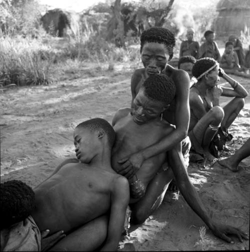 /Gunda and /Ti!kay in trance in a daytime dance, with a man helping them, group of women and children sitting in the background