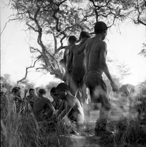 /Gunda sitting on the ground in trance at a daytime dance, with three men dancing next to him, group of people sitting in the background