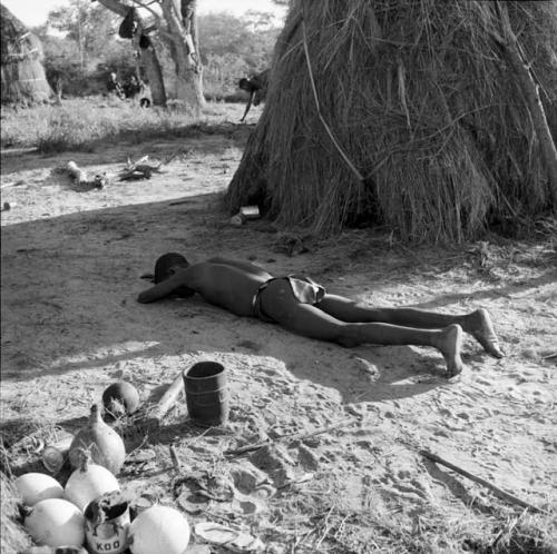 Man sleeping next to a skerm, with ostrich eggshells, mortar and pestle and a large root on the ground near him