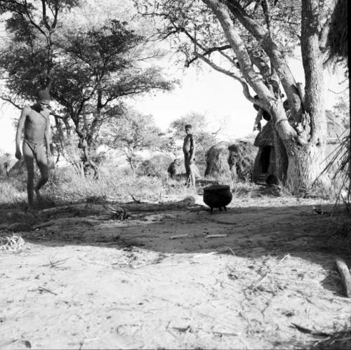 Man walking toward a fire with an iron pot on it, distant view, with another man standing in the background, skerms behind him