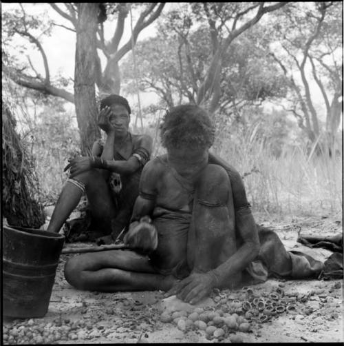 Woman sitting, cracking nuts with two stones, with another person sitting behind her