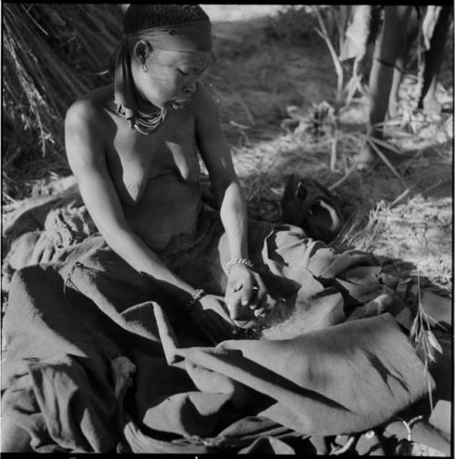 Woman sitting, pounding bone meal in her kaross with a rock