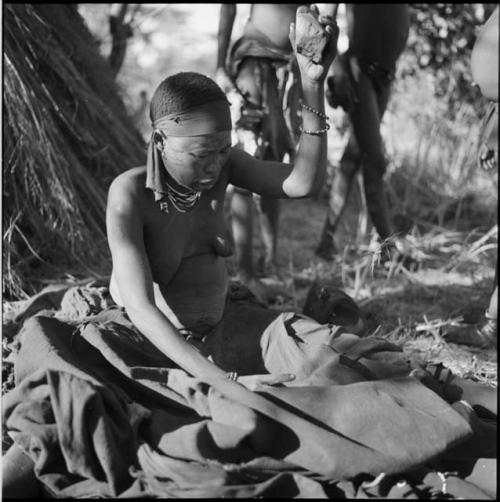 Woman sitting, pounding bone meal in her kaross with a rock