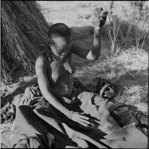 Woman sitting, pounding bone meal in her kaross with a rock