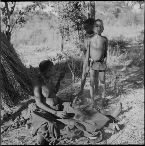 Woman sitting with her kaross wrapped around her legs, smoking a pipe, with two boys standing next to her