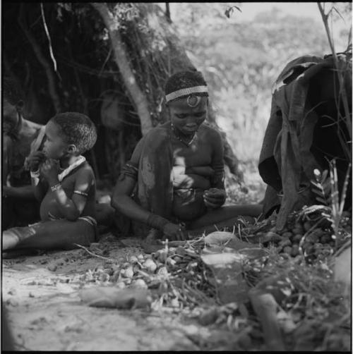 Woman looking at a nut in her hand, sitting with a pile of nuts on the ground in front of her, child next to her