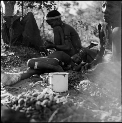 Two women sitting, cracking nuts, with a child with a bandaged foot lying on the ground between them