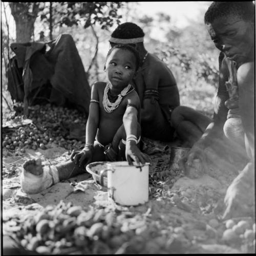 Two women sitting, cracking nuts, with a child with a bandaged foot sitting next to them, reaching for an enamel cup