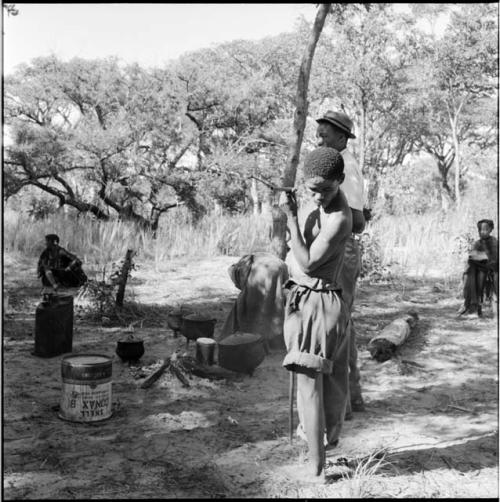 "Short /Qui" standing, holding a pipe, spitting tobacco juice on the ground, with people standing and sitting near him, jerry cans and iron pots on the ground next to him, fire burning in the background
