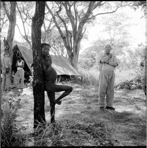 ≠Toma standing on one foot, leaning against a tree, with Laurence Marshall standing near him, two other expedition members standing in front of a tent in the background