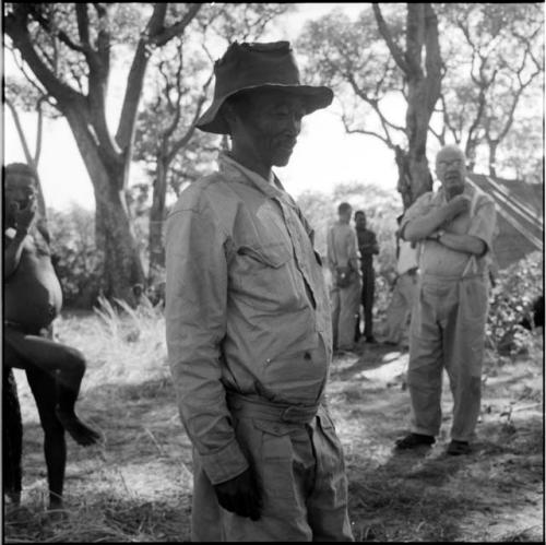 Bojo wearing Western clothing, standing, with ≠Toma and Laurence Marshall standing near him, two other expedition members standing in the background