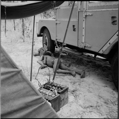 Two boys sleeping in the shade of the expedition Land Rover, with batteries on the ground near them