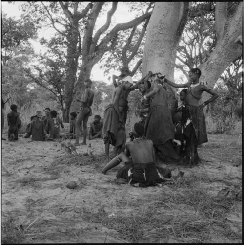 Group of women and girls playing Tcxai Djxani (a dancing game) at a tree, with a group of men and boys sitting in the background