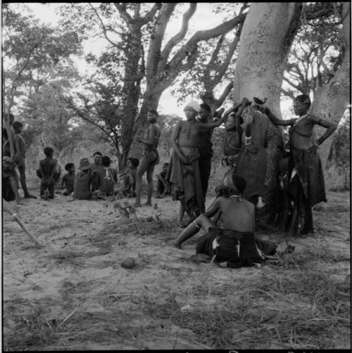 Group of women and girls playing Tcxai Djxani (a dancing game) at a tree, with a group of men and boys sitting in the background