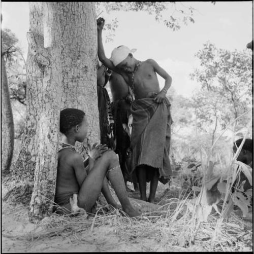 N!ai playing Tcxai Djxani (a dancing game) at a tree, with a girl sitting and clapping next to her